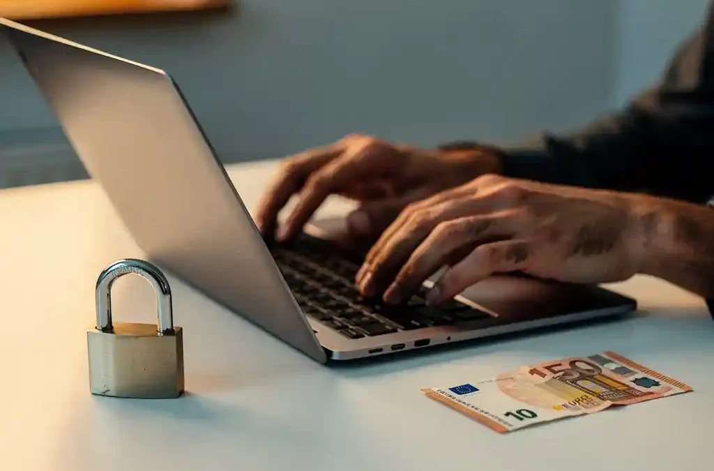 Handen van een ondernemer die een laptop opent naast een hangslot en een gevouwen eurobiljet op een bureau.