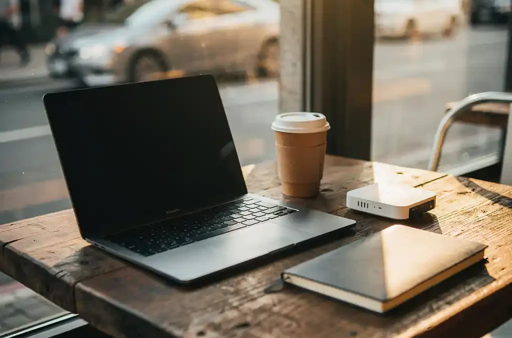 Laptop en koffie op houten cafétafel met draadloze router en notitieboek, warm middagzonlicht door stadsraam.